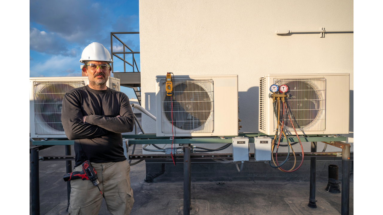 "HVAC technician standing with arms crossed on a rooftop next to two outdoor air conditioning units in Phoenix, Arizona, with gauges and tools visible."